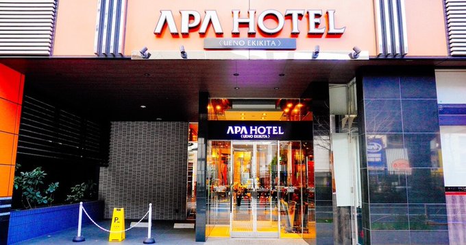 The exterior entrance of an APA Hotel (Ueno Ekikita) in Japan, with orange and black facade and glass doors.