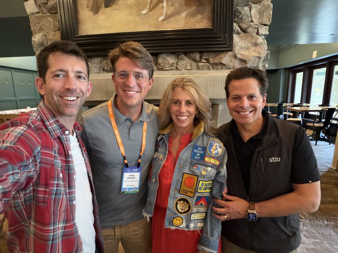 Four people pose for a photo inside a stone-accented venue, including a woman in a denim jacket adorned with various patches and a man on the right in a black "Stio" vest. The setting aligns with the caption, capturing attendees at the SALT Conference alongside Anthony Scaramucci.