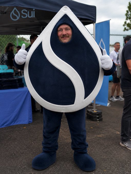 A person in a large, navy blue and white water droplet mascot costume gives two thumbs up at an outdoor event, standing beneath a 'Sui' branded tent.