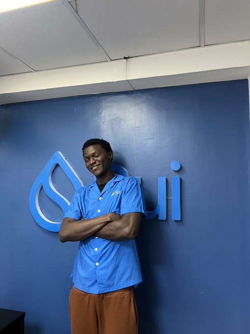 The image features a man standing against a blue wall, smiling for the camera while wearing a blue shirt with white lettering. The background of the photo shows a sign that reads "Grand rising CT I am so happy I woke up today thanks to God and being able to join morning SSM spaces with @sheisTobi_ , @doncurrent , @iamkjess and @King_Breezyy".