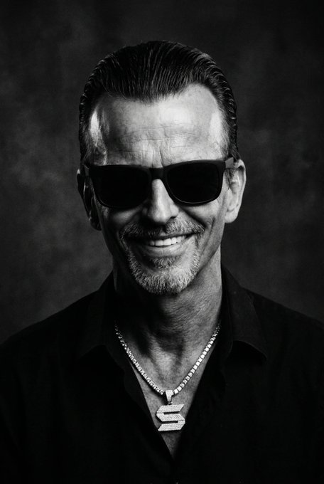 A black-and-white portrait of a smiling man with slicked-back hair wearing dark sunglasses, a black shirt, and a diamond necklace featuring an 'S' pendant.