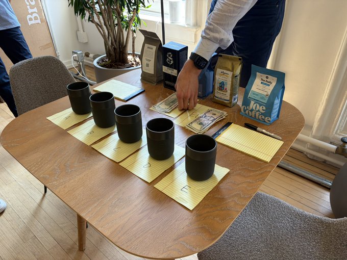A coffee tasting setup with five black cups on yellow cards numbered 1-5 on a wooden table, alongside bags of Joe Coffee and other coffee brands, with a person arranging dollar bills.