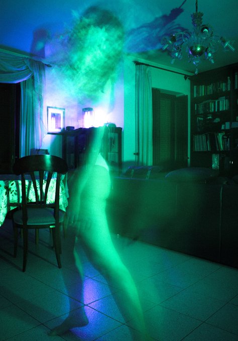The image is of a person's head standing over a chair with their body facing away from us. The background features two couches and a dining table with a lamp shining on them, creating an atmosphere reminiscent of a disco ball room or a science fiction setting. A green fog machine in the top left corner adds to the surreal ambiance of the scene, which includes text that reads: "even a corpse will dance".