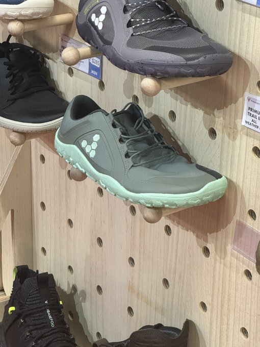 Trail running shoes displayed on a wooden pegboard wall, including a grey and mint green Vivobarefoot Primus Trail All Weather shoe.