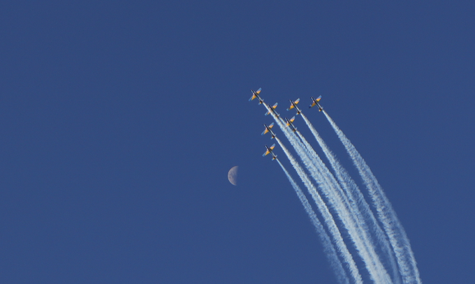 A formation of fighter jets flies in a tight diamond pattern, leaving white smoke trails across a deep blue sky beside a thin crescent moon.