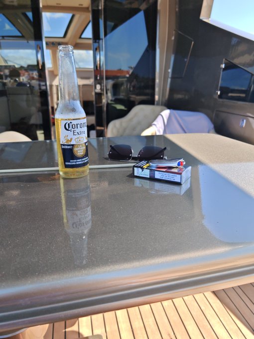 An open bottle of Corona Extra beer sits on a table next to sunglasses and a pack of cigarettes on what appears to be a boat.