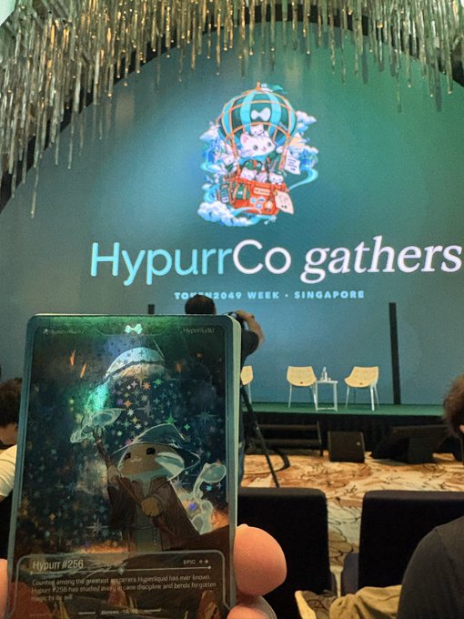 A photograph taken at a conference shows a hand holding a smartphone displaying a digital collectible card, with a presentation screen in the background reading 'HypurrCo gathers' and 'TOKEN2049 WEEK - SINGAPORE'.