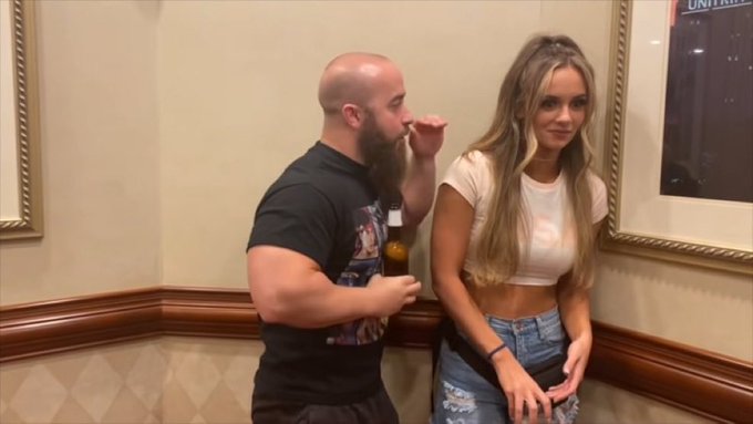 A bearded man holding a beer bottle stands close to a blonde woman in a white crop top and ripped jeans in what appears to be a hallway or lobby.