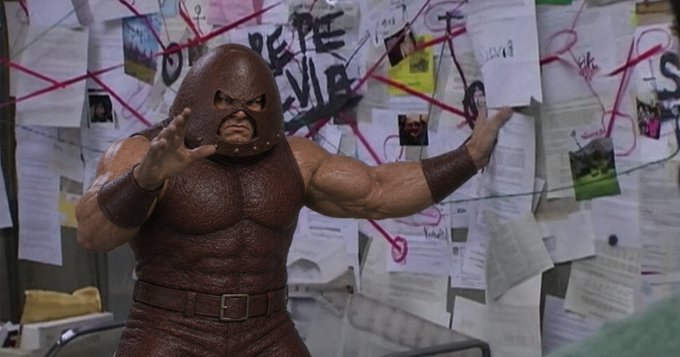 A still from a film showing a muscular man in a brown hooded costume standing in front of a bulletin board covered in papers and string, with graffiti reading 'ONE PE VIE' visible behind him.
