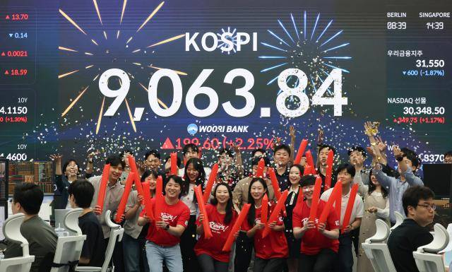 A photograph of a group of people celebrating in front of a large electronic stock board displaying the KOSPI index at 9,063.84, showing a decline of 199.60 (-2.25%), with times shown for Berlin and Singapore.