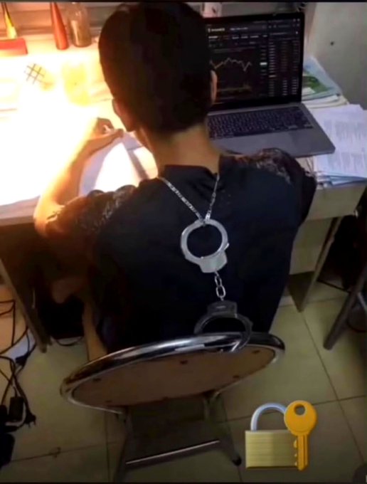 A person sits at a desk facing a laptop displaying charts, wearing handcuffs connected by a chain to a metal ring, with an unlocked padlock emoji in the bottom right corner.