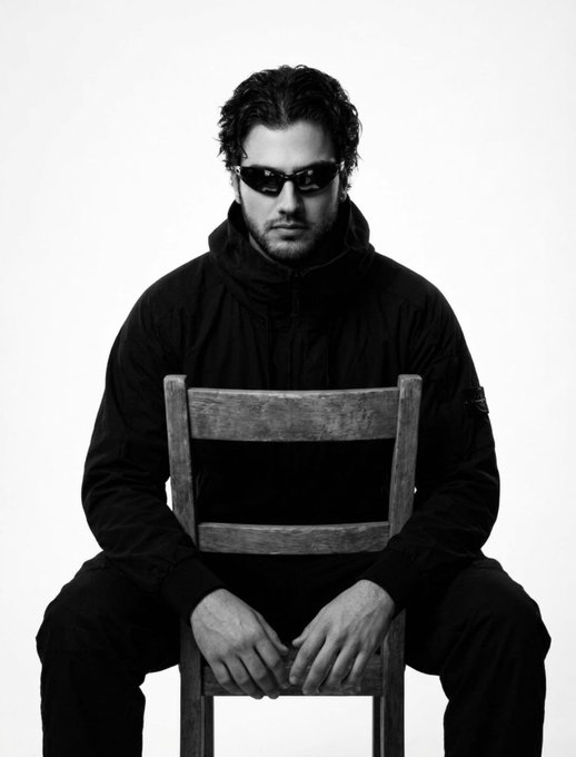 A man wearing dark sunglasses and a black jacket sits on a wooden chair in this black-and-white photo, his hands clasped in front of him.