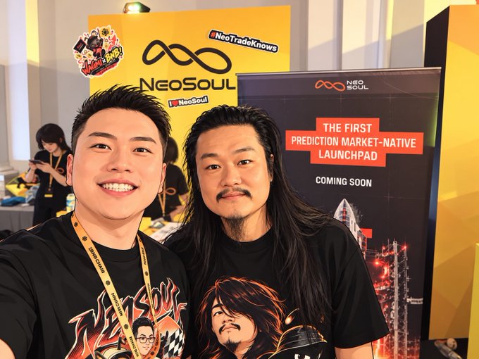 Two men smile for the camera at what appears to be a crypto conference, standing in front of a 'NeoSoul' booth advertising themselves as 'THE FIRST PREDICTION MARKET-NATIVE LAUNCHPAD COMING SOON'.