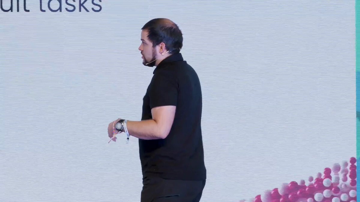 The image shows a man standing in front of a white wall that has pink and purple circles on it, possibly representing a logo or design element related to the post about Flares smart accounts. The man appears to be giving a presentation or speech at an event, as indicated by his attire - he is wearing a black shirt and a headset around his neck.