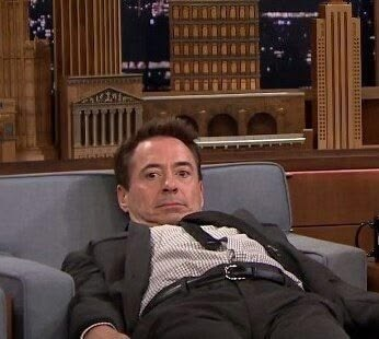A photo of actor Robert Downey Jr. appearing on a late-night talk show set, slouched in a chair and wearing a dark suit with suspenders.