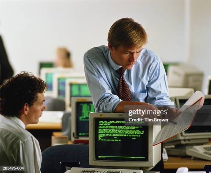 A man in a blue shirt and tie leans over a computer monitor displaying green text on a black screen, scrutinizing a spreadsheet held in his hand while another man at a nearby desk looks on. The retro tech setup and serious expressions evoke a Wall Street trading floor, fitting the caption's jab at Robinhood locking out users during the GameStop short squeeze.