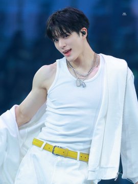A young male K-pop performer with dark hair wearing a white tank top, yellow belt, and white jacket, accessorized with a silver necklace.