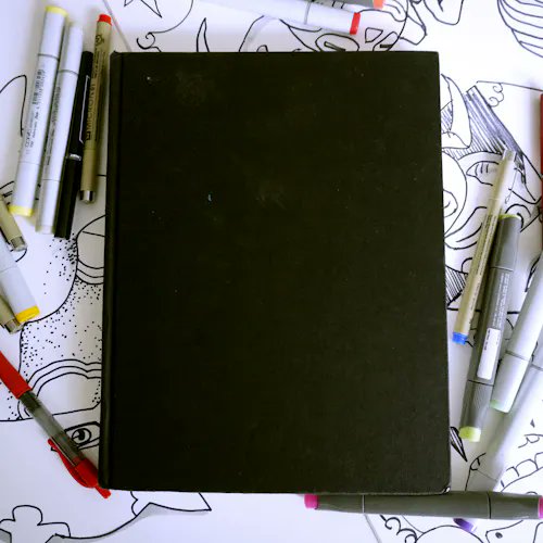 A black sketchbook lies closed on a surface covered with line drawings of faces, surrounded by various markers and pens in white, gray, and red.