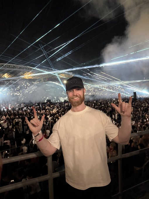 A bearded man in a beige t-shirt and black cap makes rock hand gestures while posing in front of a large concert crowd illuminated by sweeping laser lights and stage smoke.