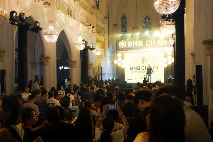 The image shows a large audience seated and standing in an ornate hall featuring grand chandeliers and arched windows, attentively facing a stage prominently displaying "BNB CHAIN SUPER MEETUP." A speaker stands on stage, with "BNB CHAIN" branding visible in golden yellow logos and screens, perfectly capturing the moment described—crowds ready and the stage set for an evening of insights.