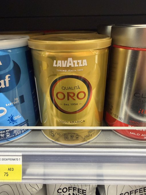 A supermarket shelf displaying a gold Lavazza Qualità Oro coffee tin alongside other coffee products, with a yellow price tag showing 'AED 75'.
