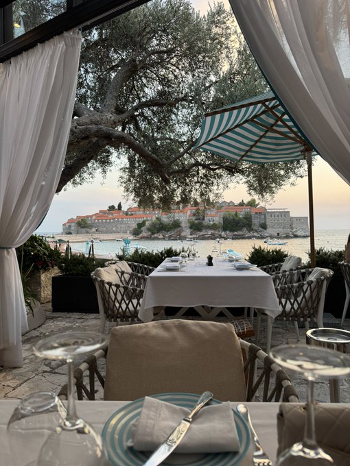 An elegantly set dining table overlooks a stunning coastal view of a historic town across the bay, framed by white curtains and a striped umbrella. The scene captures a luxurious seaside escape, suggesting the lavish lifestyle one enjoys after consistently winning on @Stake.