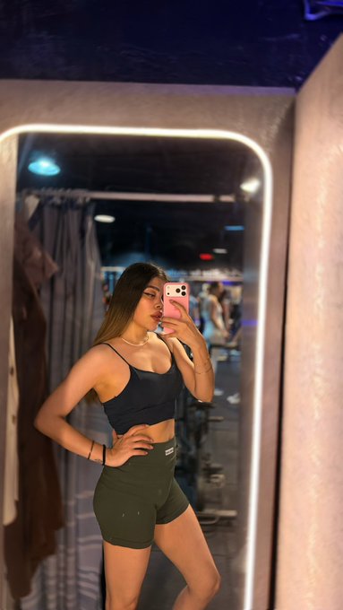 A woman taking a mirror selfie with a pink-cased phone, wearing a black crop top and olive green athletic shorts, in what appears to be a gym or locker room.