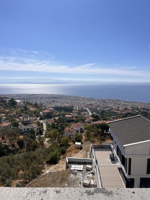 A scenic elevated view overlooking a coastal town and a vast ocean under a clear blue sky, with houses and a modern building with a terrace in the foreground.