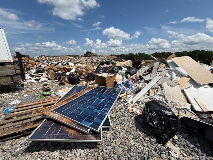 A photograph of a landfill or dumping site showing scattered debris, cardboard, black trash bags, and discarded solar panels on a rocky surface under a partly cloudy sky.