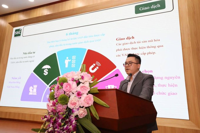 The image features a man standing at a podium, giving a speech or presentation. The man appears to be wearing glasses and has a serious expression as he speaks into a microphone. Behind him, there's a large screen displaying information related to his topic of discussion. The setting seems to be an indoor event space with wooden walls, providing a professional atmosphere for the gathering.