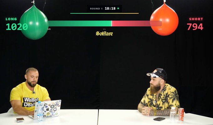 Two men sit at a white table during a competition titled 'Solfare' with balloons above them showing 'LONG 1020' and 'SHORT 794', and a timer reading 'ROUND 1 18:18'.