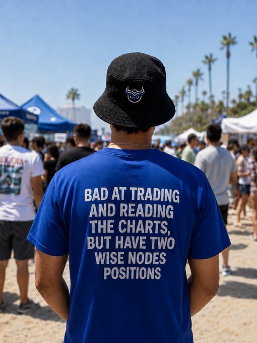 A person viewed from behind wears a black bucket hat with a bull logo and a blue t-shirt reading 'BAD AT TRADING AND READING THE CHARTS, BUT HAVE TWO WISE NODES POSITIONS' at a beachside event.
