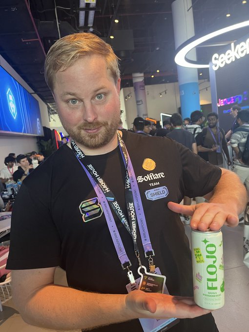 A man with a beard wearing a black 'Solflare TEAM' t-shirt and a Hackathon lanyard holds a can of 'FLOJO' at what appears to be a convention or event.