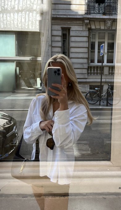 A blonde woman in an oversized white button-up shirt takes a mirror selfie with her iPhone, accessorized with sunglasses and a crossbody bag, capturing a stylish Parisian street scene with classic European architecture, a parked car, and a bicycle in the background—perfectly matching her casual and chic "gm babies" morning greeting.