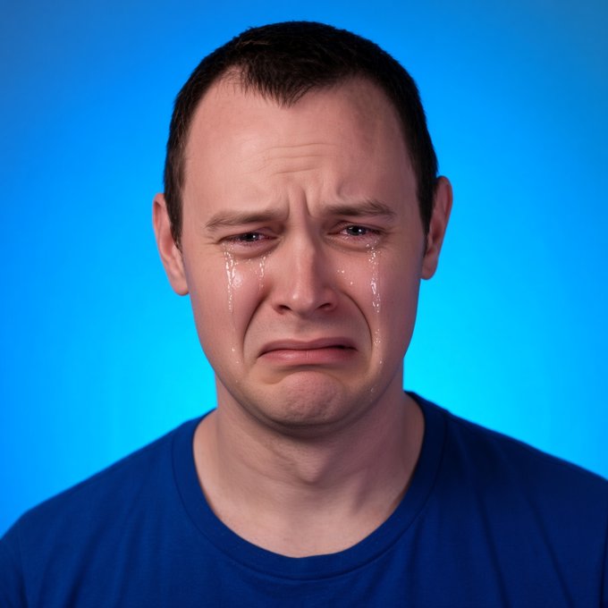 A man with short dark hair and a blue t-shirt is crying with tears streaming down his face against a bright blue background.