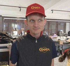 A smiling man wearing a red "CAMBRIA" cap and a black polo shirt featuring the Cambria logo stands in what appears to be a clothing warehouse or workshop, with folded garments stacks and tables visible in the background. The image ties to the post by visually representing the Cambria team behind the web3 gaming project celebrated for being officially recognized by Ethereum, marking a milestone in the project's evolution from early development to its Duel Arena era.