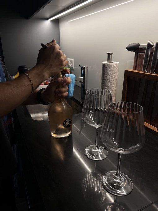 A person uses a corkscrew to open a bottle of white wine in a kitchen setting, with two empty wine glasses and a knife block on the counter.