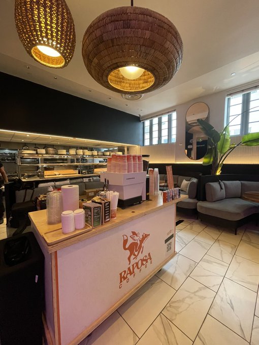 An interior shot of a Raposa Coffee shop featuring a wooden counter with the brand's fox logo, woven pendant lights, and a view into the kitchen area.