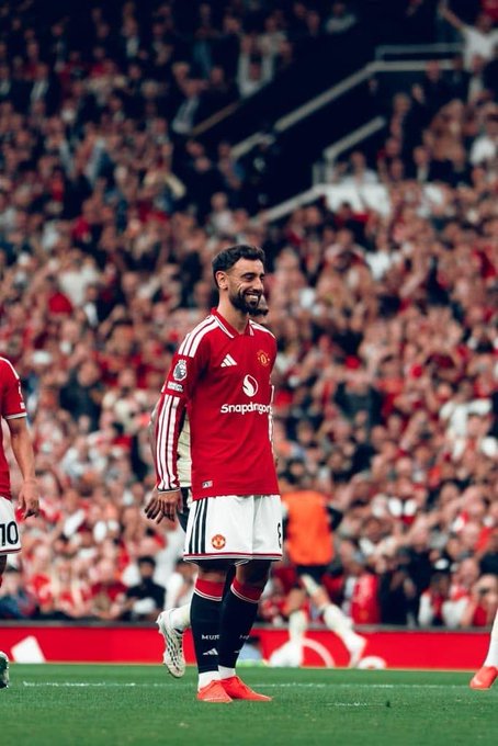 A photo of Manchester United player Bruno Fernandes wearing the team's red home kit with the Snapdragon sponsor logo, smiling on the pitch with a crowd in the background.