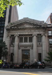 An exterior view of a classical-style stone building with 'NATIONAL COMMERCIAL BANK' engraved on the pediment, featuring columns and arched doorways.