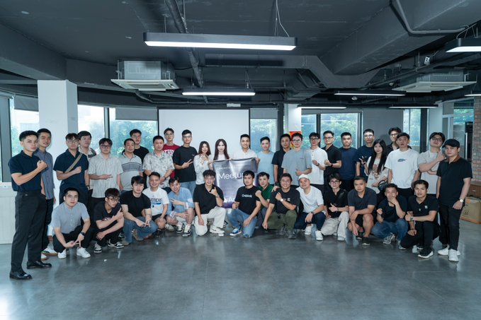 A group photo of approximately 30 people posing together in a modern office or classroom space, with a 'MetaUp' banner visible in the center.