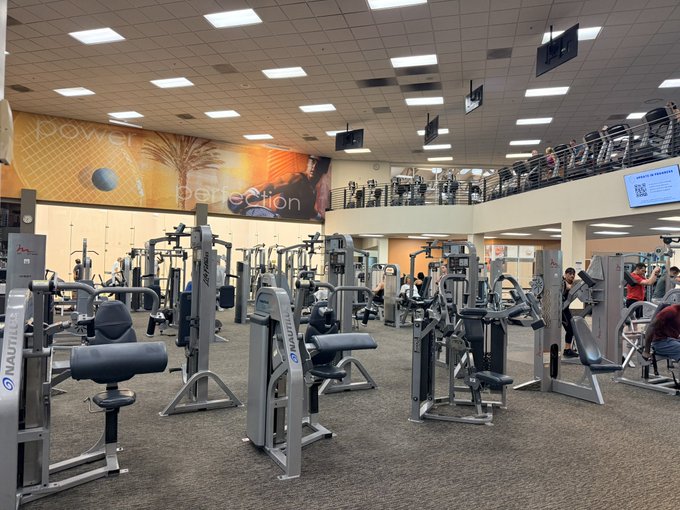 A well-equipped fitness center with Nautilus weight machines is shown under bright ceiling lights, featuring a large mural with the words "Power" and "Perfection" on the back wall, with several gym-goers working out among the equipment, aligning with the early morning gym routine mentioned in the post.