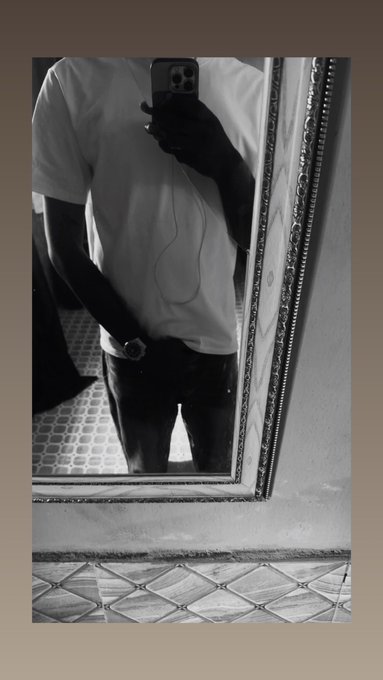 A black and white mirror selfie of a person wearing a white t-shirt and dark jeans, with their face hidden behind a smartphone.