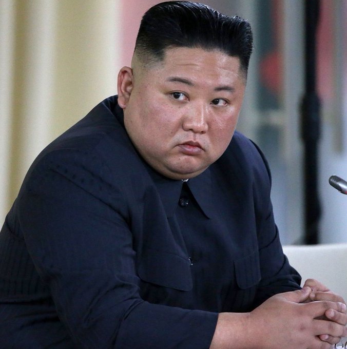 A photograph of Kim Jong Un wearing a dark navy Mao-style suit, seated with his hands clasped together.