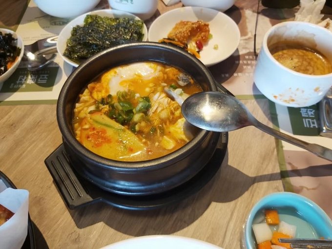 A bubbling Korean stew in a black stone pot sits at the center of a wooden table, surrounded by side dishes including seaweed, fried items, and what appears to be instant noodles in a paper cup, with a small salad visible in the foreground. The post discusses late-night eating while watching football matches and managing sleep with a sleep-tracking app, suggesting this is a late-night meal accompanying the author's soccer viewing.
