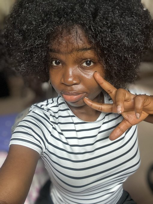 A close-up selfie of a young woman with natural curly hair wearing a black and white striped t-shirt, making a hand gesture near her face.