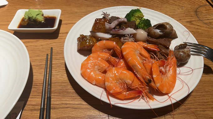 A plate of cooked shrimp with vegetables and a side of wasabi sits on a wooden table alongside chopsticks and a fork, likely depicting the meal shared between the two travelers mentioned in the post.