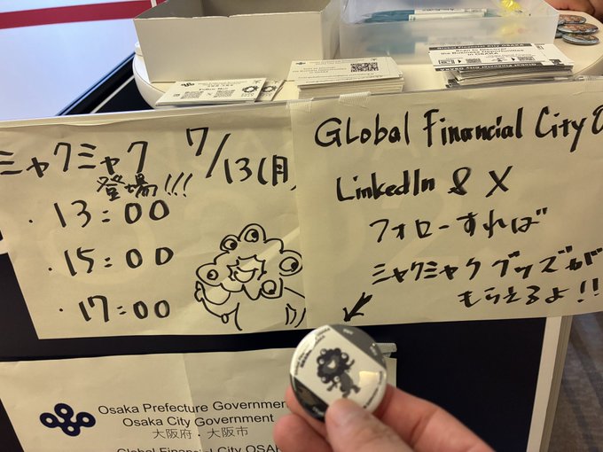 A hand holds a round "Myaku Myaku" badge in front of a handwritten sign at the Osaka City Government booth, which lists times (13:00, 15:00, 17:00) and promotes the Global Financial City, LinkedIn & X event featuring Minakata and Tonomura ("ミナカタ / トノムラ") at WebX2026.