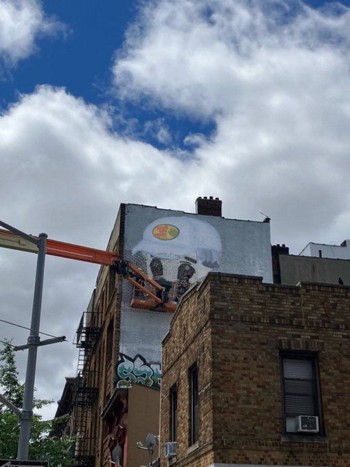 A cherry picker lifts an artist working on a large "Milady" mural on the side of a brick building, with the iconic meme-style face visible beneath partly cloudy skies. The graffiti-style lettering "S27" appears in teal near the bottom of the wall.