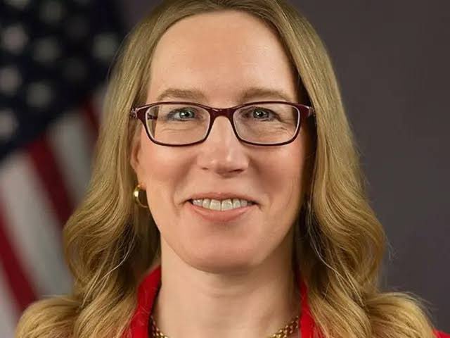 A professional headshot of a woman with long blonde hair wearing burgundy-framed glasses, with an American flag visible in the background, suggesting an official government photo.